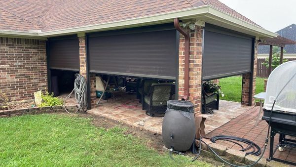 Brown roll-down shutters cover an outdoor patio. The brick patio has a rain barrel, furniture, and is surrounded by a lawn.