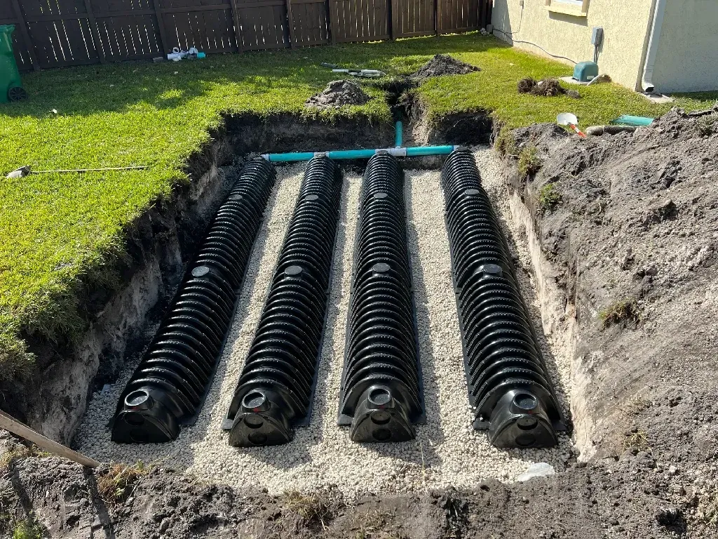 Three black corrugated drainage pipes laid in a trench beside a house, with soil and grass around them