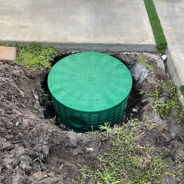 Green utility access lid in a ground pit beside a sidewalk and grass.