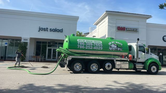 Green septic tanker truck parked outside storefronts under a clear sky