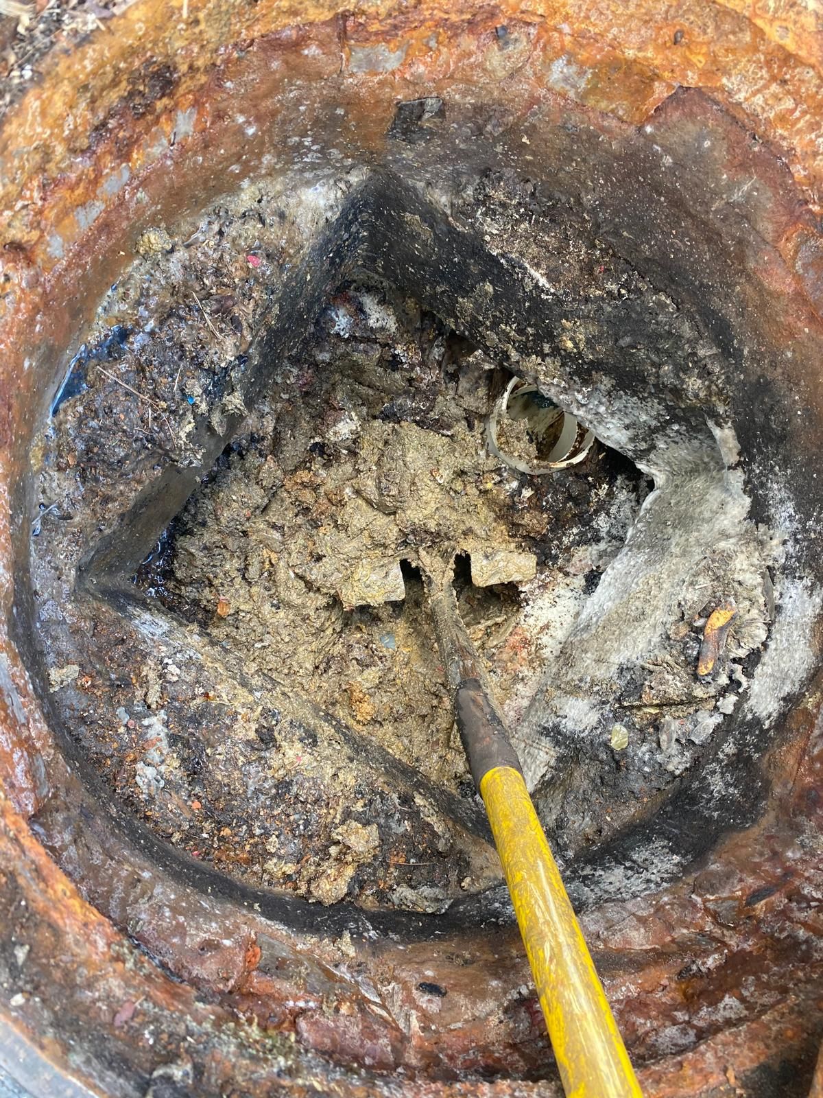 Close-up of a rusted pipe opening with debris inside and a yellow-handled tool scraping it out