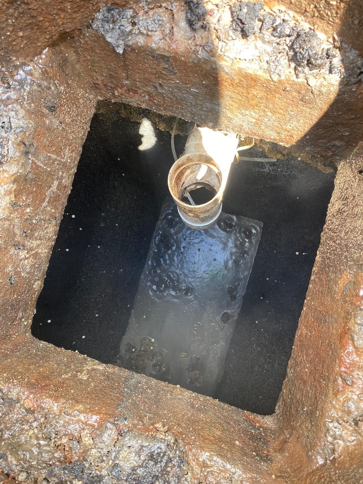 Open square manhole with a pipe opening and dark water inside, surrounded by rusty metal.