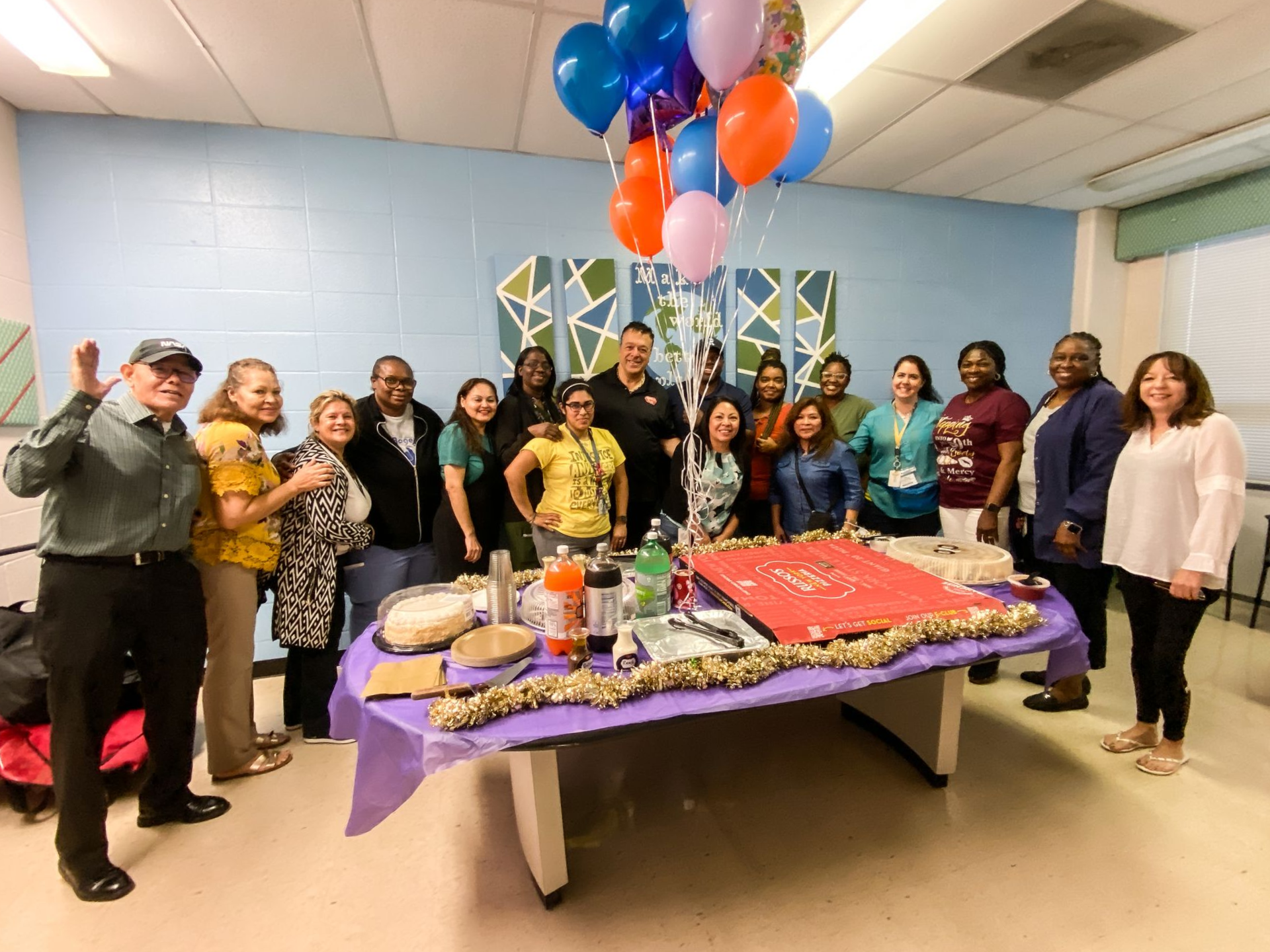 Group of people gathered around a decorated table with food and balloons; celebration in a brightly lit room.