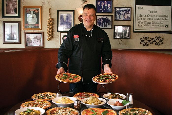 Chef holding two pizzas, surrounded by many dishes on a table in a restaurant. The chef is smiling, the setting is warm.