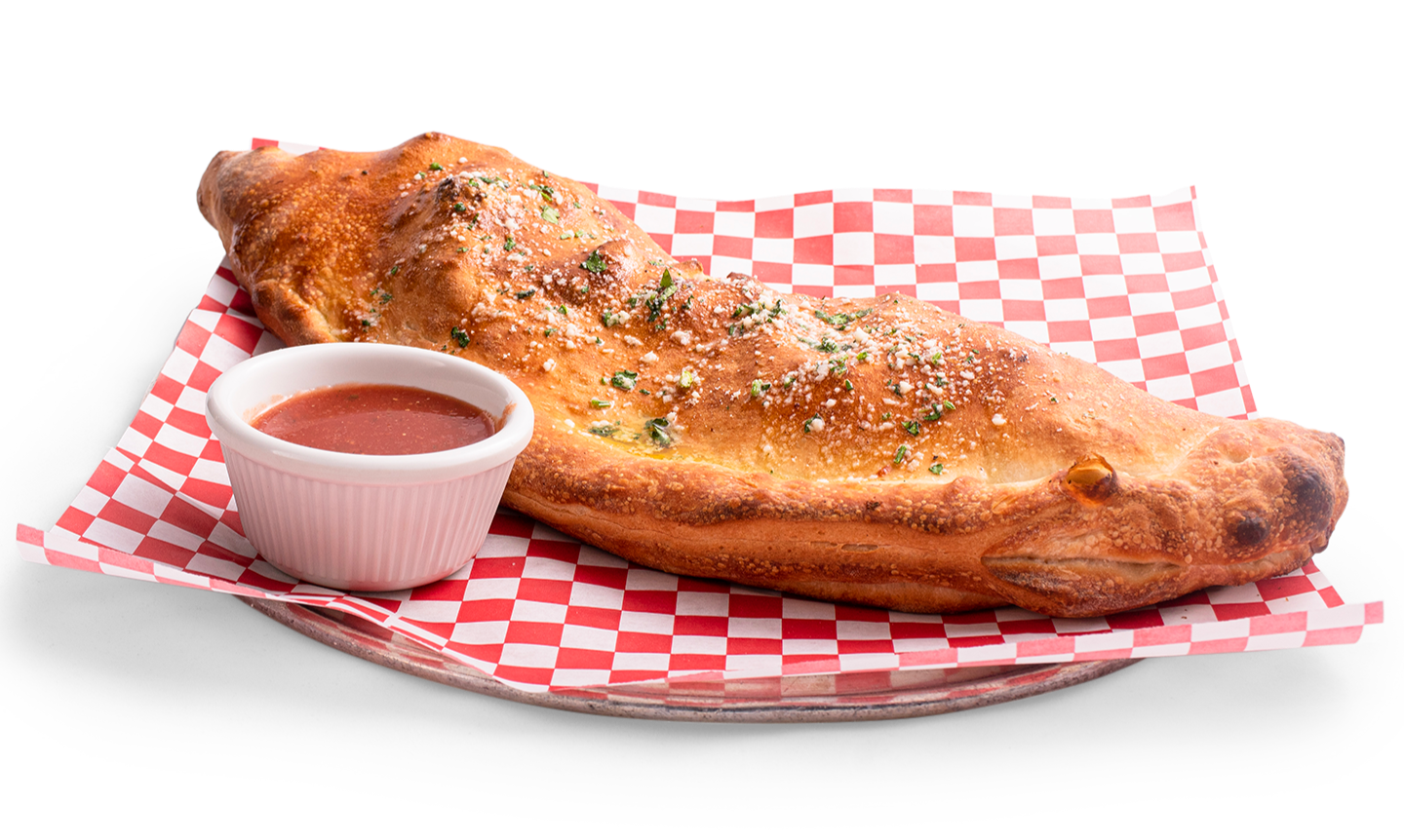 A golden-brown calzone with herbs on a red and white checkered paper liner next to a small cup of red dipping sauce.