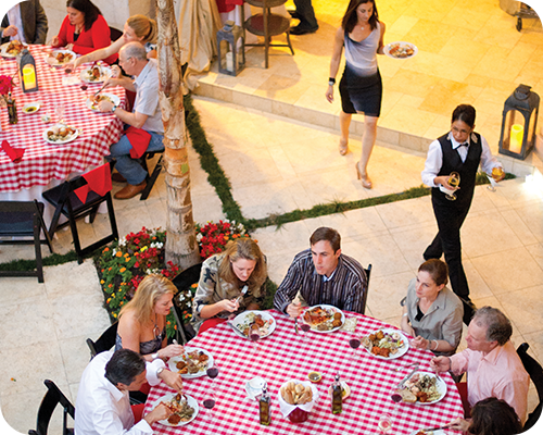 Outdoor dining scene: Several people at red-checkered tables, eating. Waiter serves.
