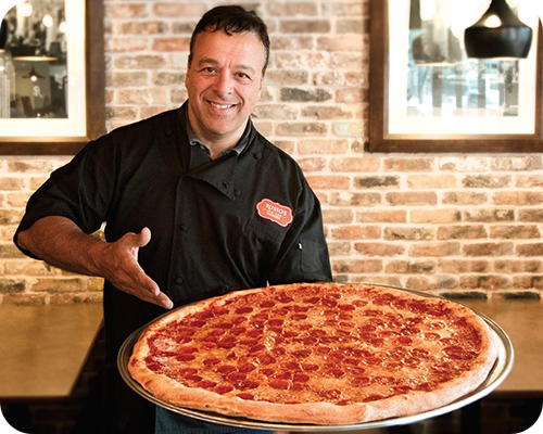 Chef holding a large pepperoni pizza, smiling, standing in a restaurant setting with brick wall.
