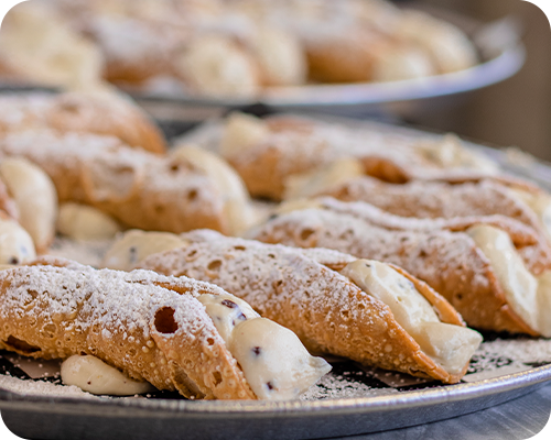 Cannoli pastries, dusted with powdered sugar, arranged on metal trays.