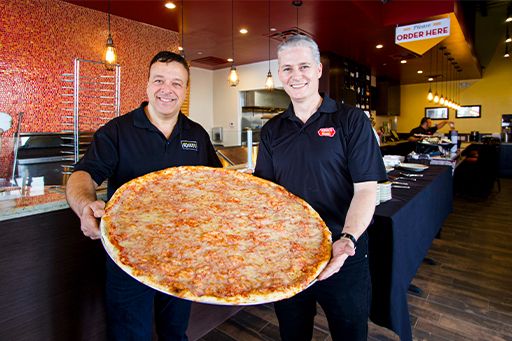 Two men in black shirts hold a giant pizza in a restaurant setting.