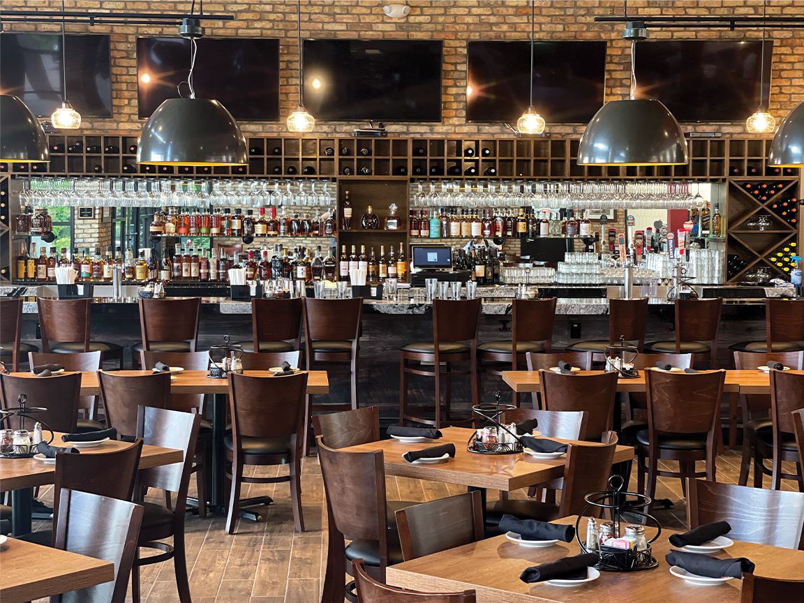 Upscale restaurant interior with wooden tables, dark chairs, brick walls, and bar shelves lit by hanging lamps