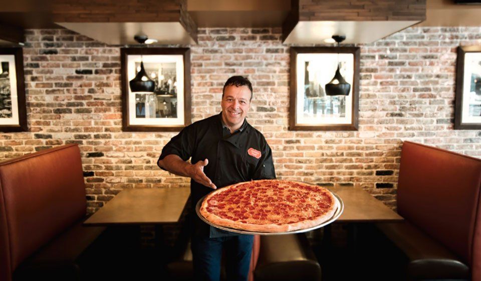 Chef Anthony Russo holding a large pizza in a brick-walled restaurant booth, smiling at the camera.