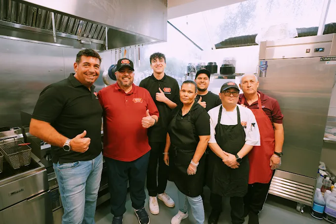Group of five smiling people posing together in a commercial kitchen, wearing aprons and caps.