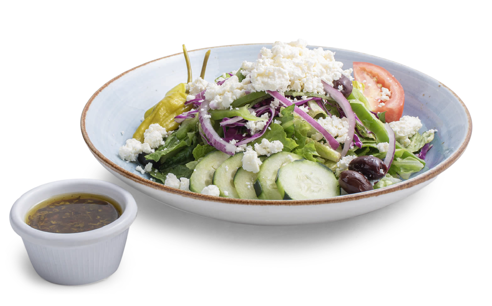 A Greek salad in a shallow blue bowl with a side of dressing, featuring cucumbers, tomatoes, feta, and kalamata olives.