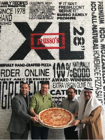 Four men holding a large pizza in front of a Russo's XL Pizza sign.