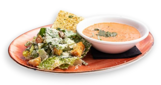 A meal on a plate featuring a Caesar salad with croutons, a piece of flatbread, and a bowl of creamy tomato soup.