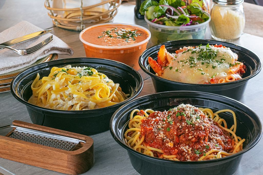 Various pasta dishes in black bowls with a side salad and soup, ready for takeout.