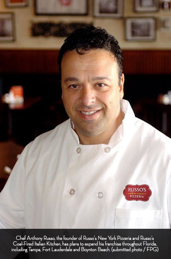Chef Anthony Russo smiling, wearing a white chef's coat with a logo pin. Restaurant interior background.