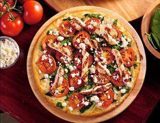 Pizza topped with sliced tomatoes, feta cheese, and greens, on a wooden board, with fresh tomatoes and cheese in the background.