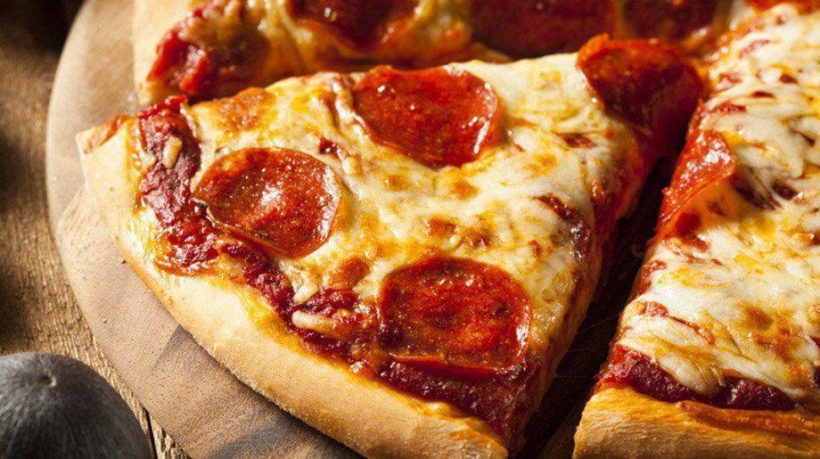 Pepperoni pizza slice on a wooden board with melted cheese, visible sauce, and more pizza in the background.
