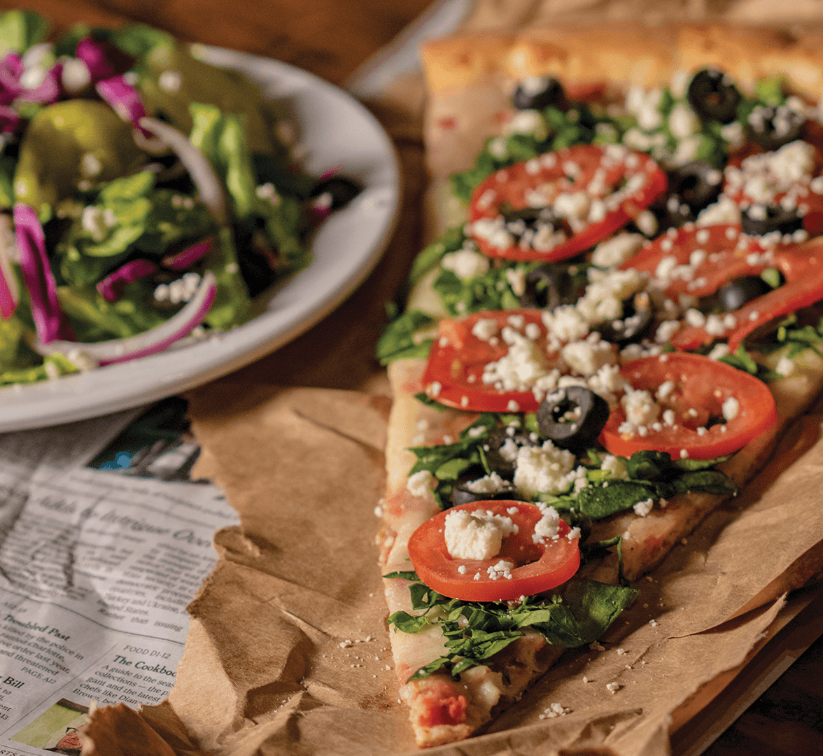 Slice of vegetable pizza on brown paper beside a green salad bowl