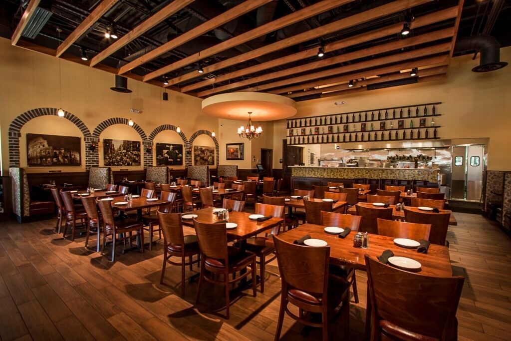An empty restaurant interior featuring wooden tables, chairs, arched wall decorations, and an open kitchen in the background.