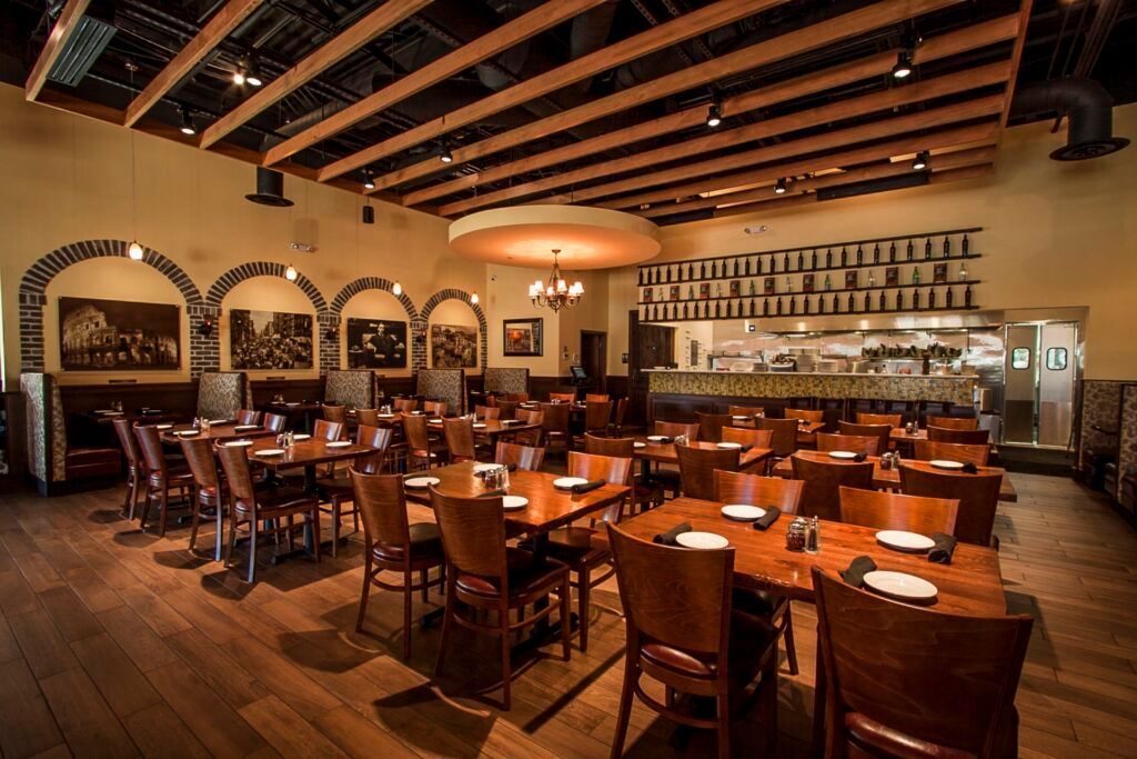 Interior of a restaurant with wooden tables and chairs. A large open kitchen is visible in the back.
