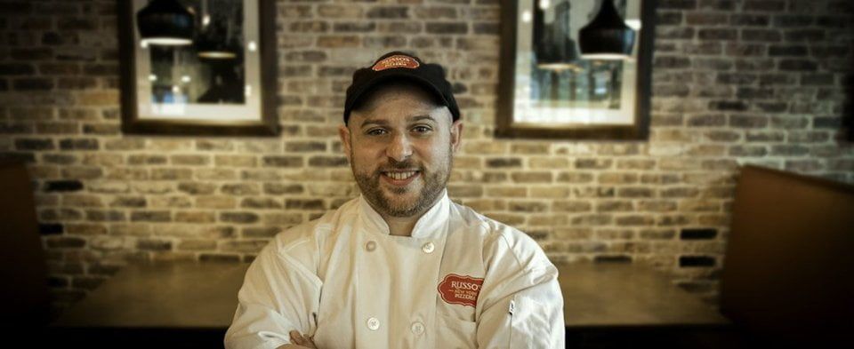 A smiling chef in a white coat and hat, arms crossed, stands in front of a brick wall.