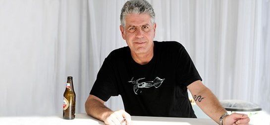 Anthony Bourdain sitting at a table, wearing a black t-shirt, with a beer bottle on the left and a white curtain backdrop.