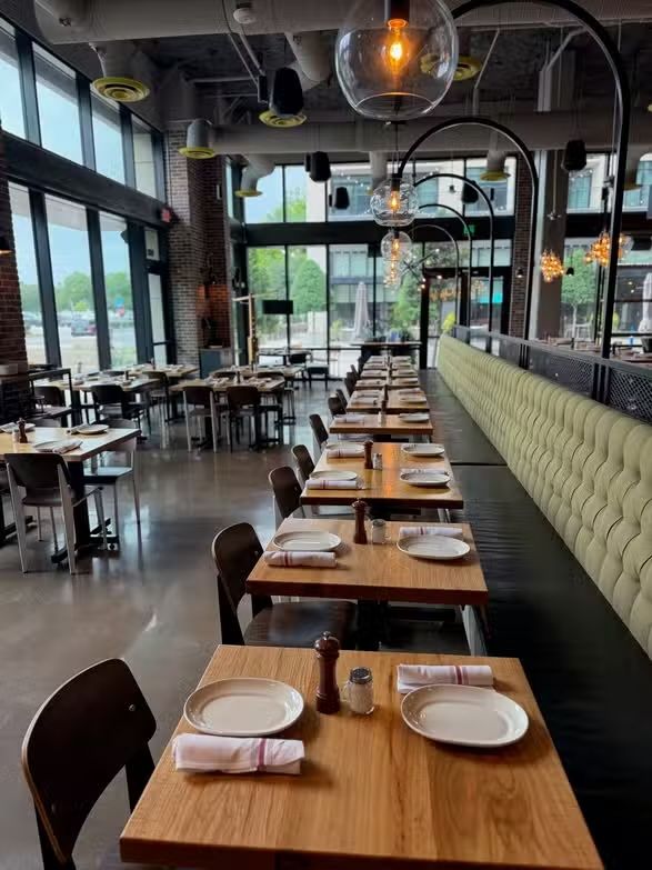 Empty modern restaurant dining room with wooden tables, black chairs, large windows, and warm hanging lights