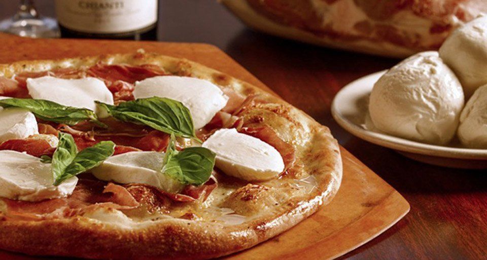 Pizza with mozzarella, basil, and prosciutto on a wooden board, with a bottle of wine and rolls.