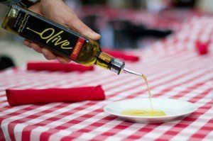 A hand pouring olive oil from a bottle onto a small white plate, set on a red and white checkered tablecloth.