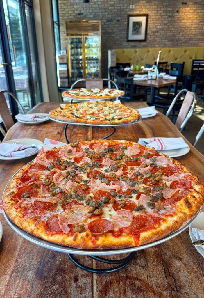 Pizza on a wooden table in a brick-walled restaurant, with a large pepperoni pizza in the foreground and pizzas behind.