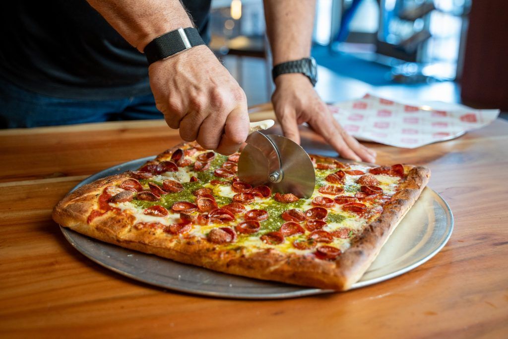 Person cutting a square pizza with a pizza cutter on a metal tray, wooden table.