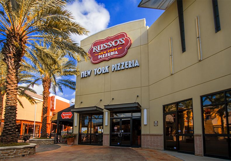 Russo's New York Pizzeria storefront. Beige building with red logo, black trim, large windows, and palm trees.