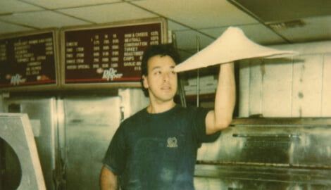 Chef Anthony Russo in dark shirt tossing pizza dough in a restaurant kitchen under menu boards