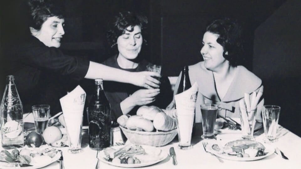 Three people sharing drinks and a meal at a dinner table, smiling and chatting.