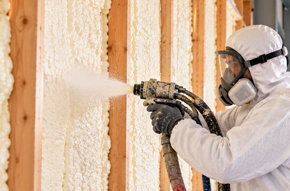 Skilled spray foam insulation technician wearing full white protective gear, gloves, and a respirator applying insulation between wooden wall studs while another technician operates professional spray foam equipment in Fall River, Massachusetts.