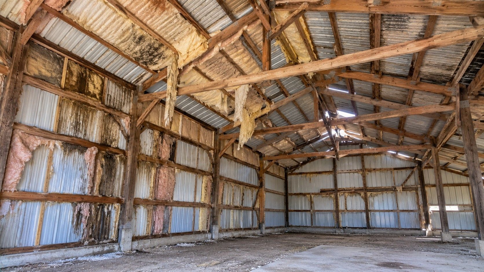 Old pole barn with damaged fiberglass insulation, exposed roof framing, moisture stains, and poorly insulated walls requiring professional insulation improvements.