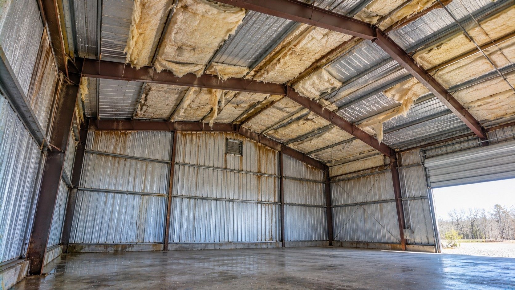 Poorly insulated metal building with damaged fiberglass insulation, exposed corrugated metal panels, condensation, and gaps requiring professional spray foam insulation.
