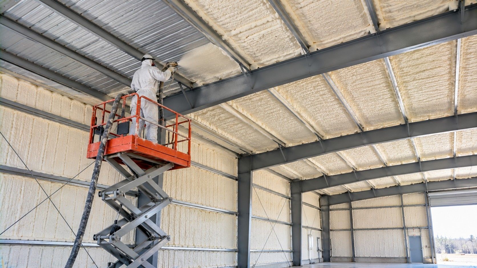 Professional insulation technician on an elevated scissor lift applying closed-cell spray foam insulation to the metal roof of a commercial building.