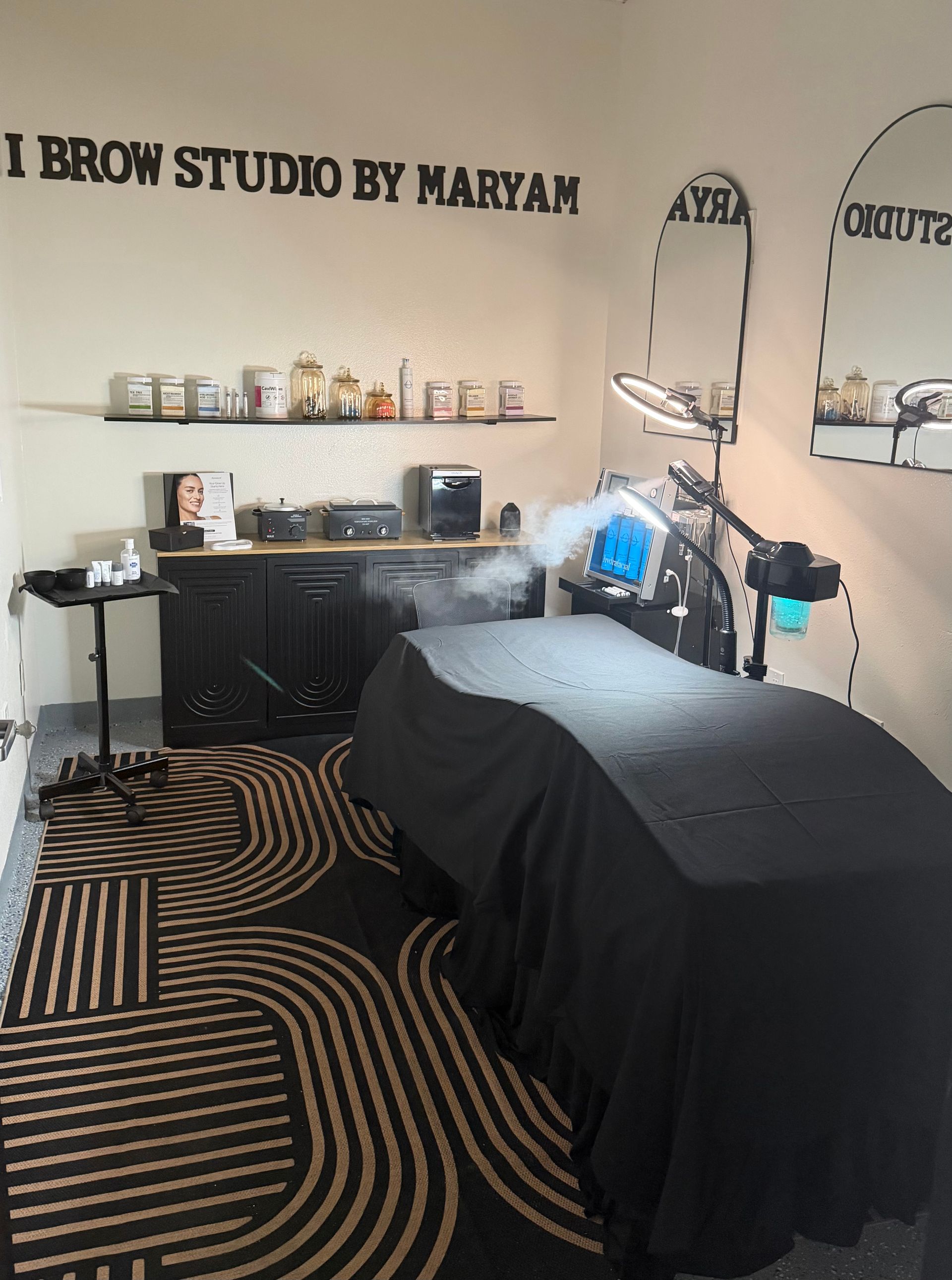 Beauty studio treatment room with black bed, shelving, desk, and patterned rug.
