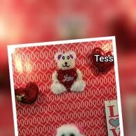 A teddy bear on a red patterned background with a white border and the word “Tess” near the top right