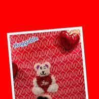 Small white teddy bear in a red heart-patterned card with a red heart balloon and “Huggable” text.