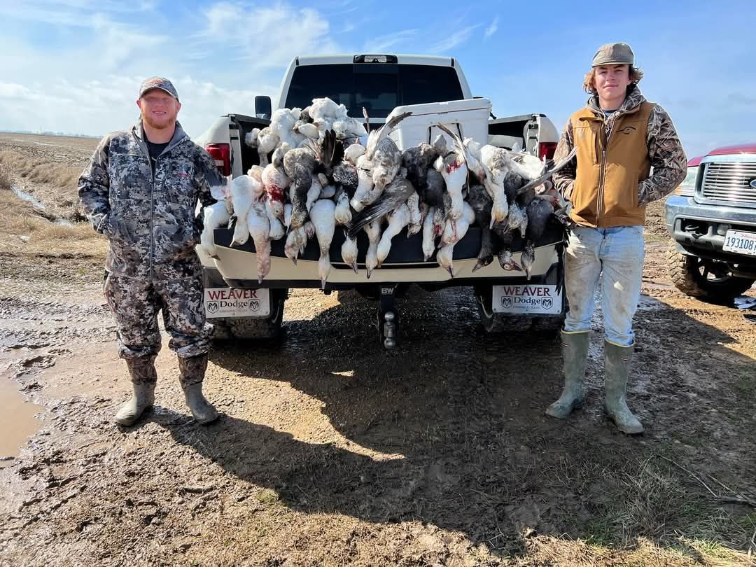 Two men pose with a truck bed full of ducks in a muddy field.