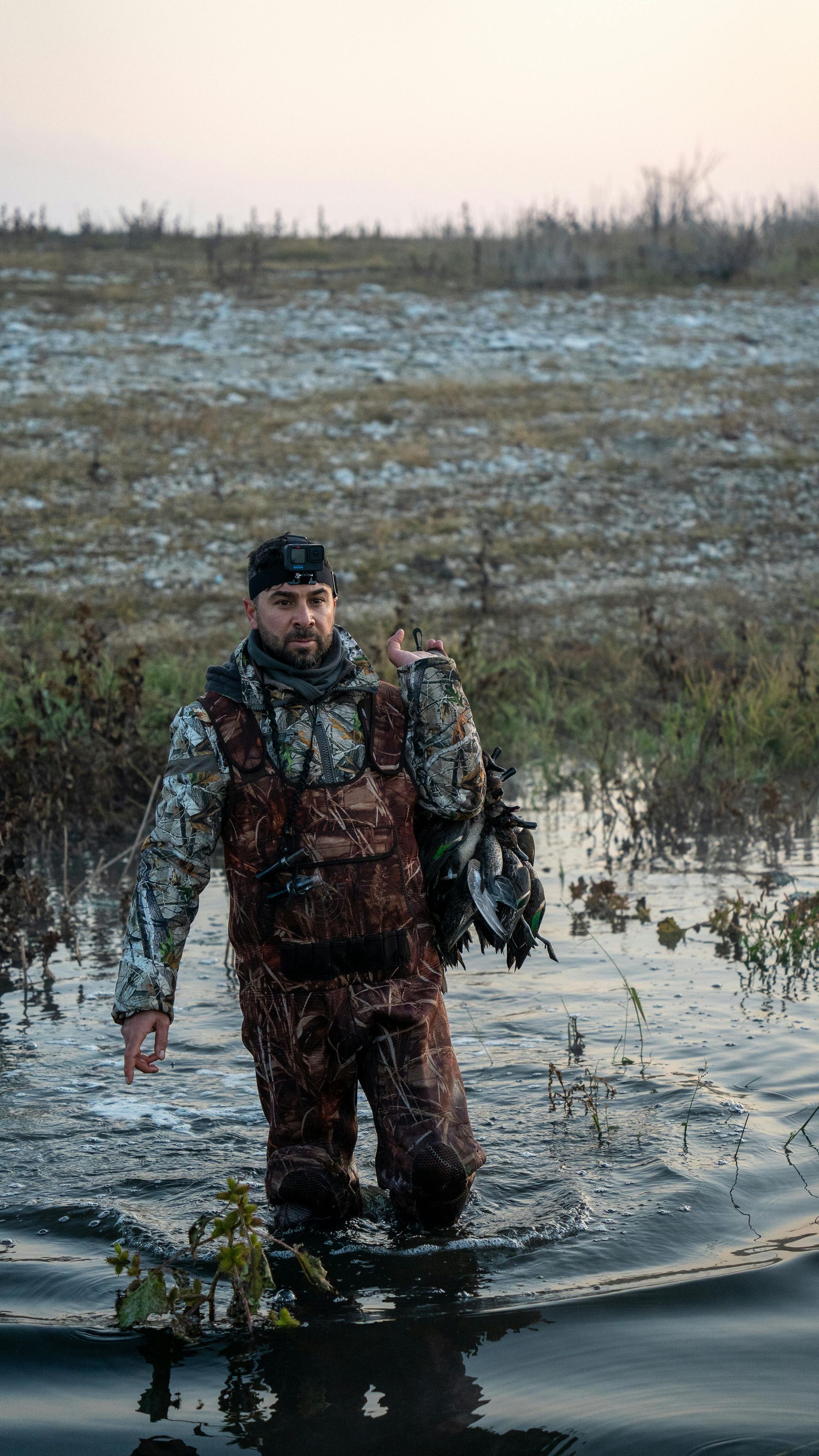Man standing in a muddy marsh, wearing camouflage and holding gear, with a cold wetland in the background