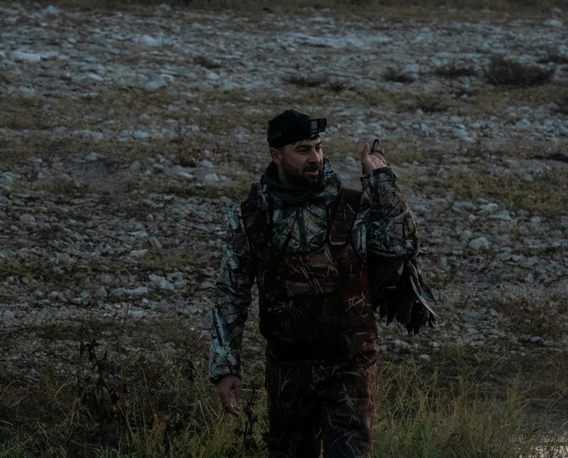 Man in camouflage carries a bird in a field at dusk.