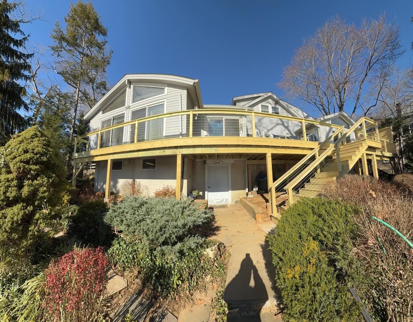 House with a large wooden deck and stairs, viewed from a slightly uphill, overgrown path, on a sunny day.