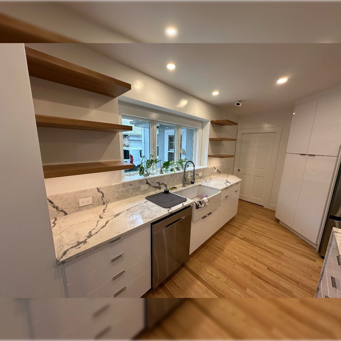 Modern kitchen with white cabinets, wood shelves, marble counters, sink, and a window over the counter