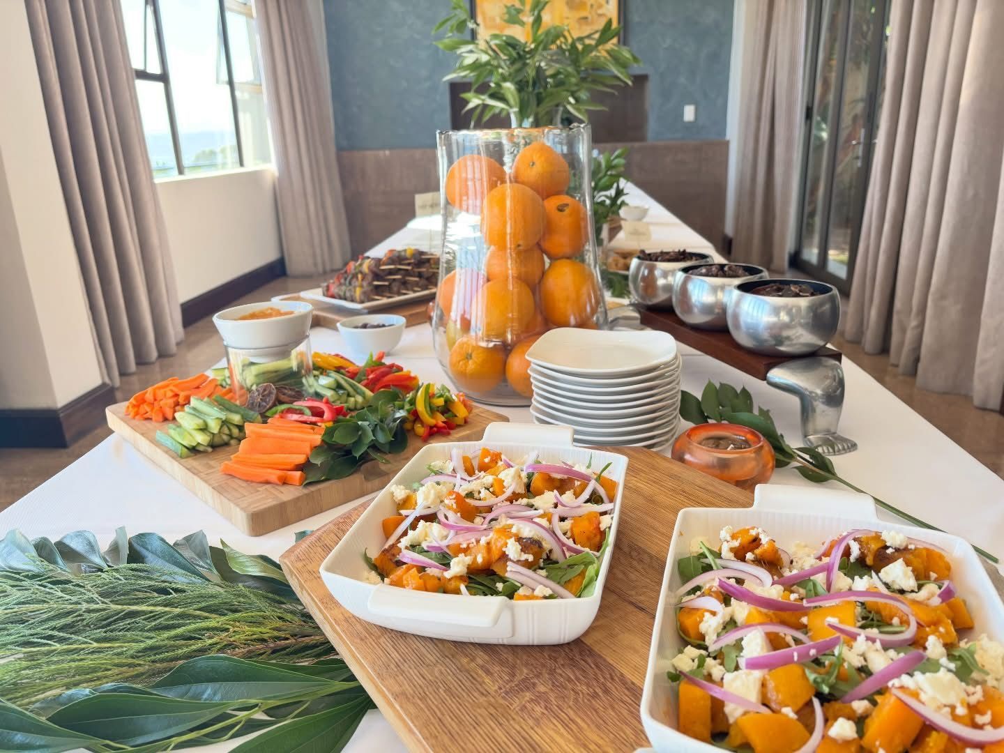 A buffet table features fresh vegetable crudités, pumpkin and onion salads, and a large central vase filled with oranges.