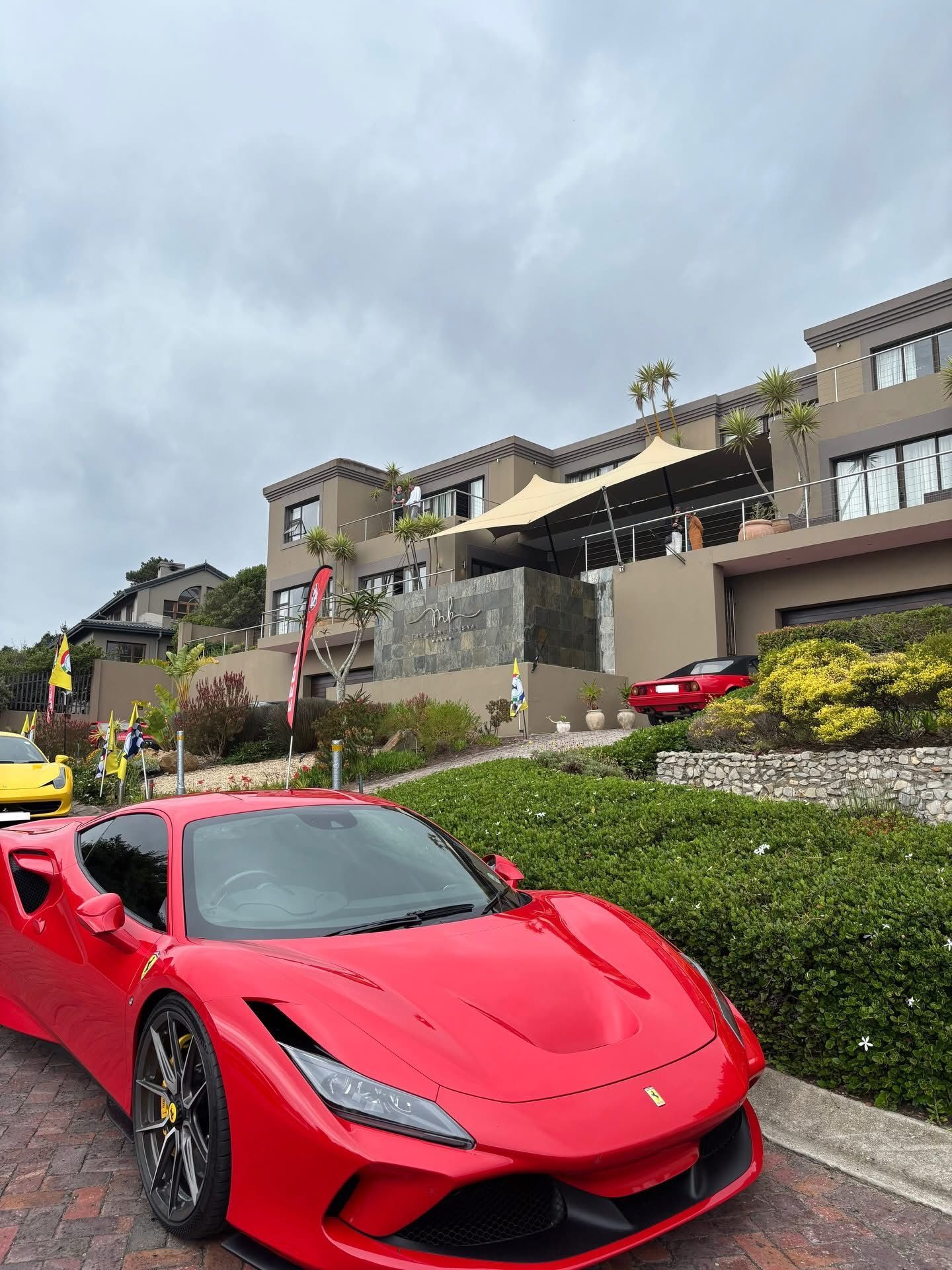 A vibrant red Ferrari sports car parked on a paved driveway in front of a modern, multi-story Mediterranean-style home.